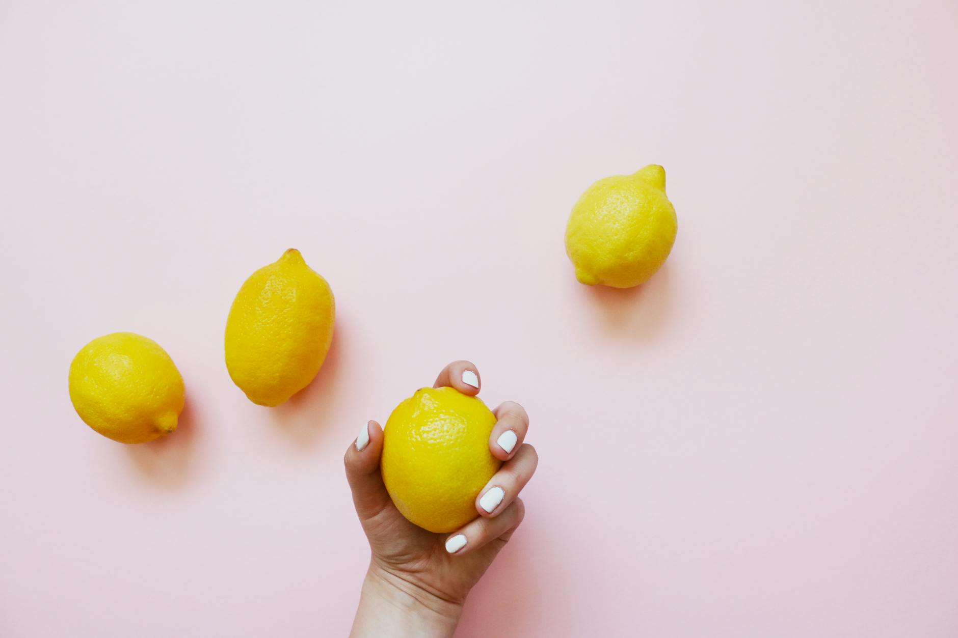 Une main tenant un citron frais sur fond rose pastel entouré de trois autres citrons