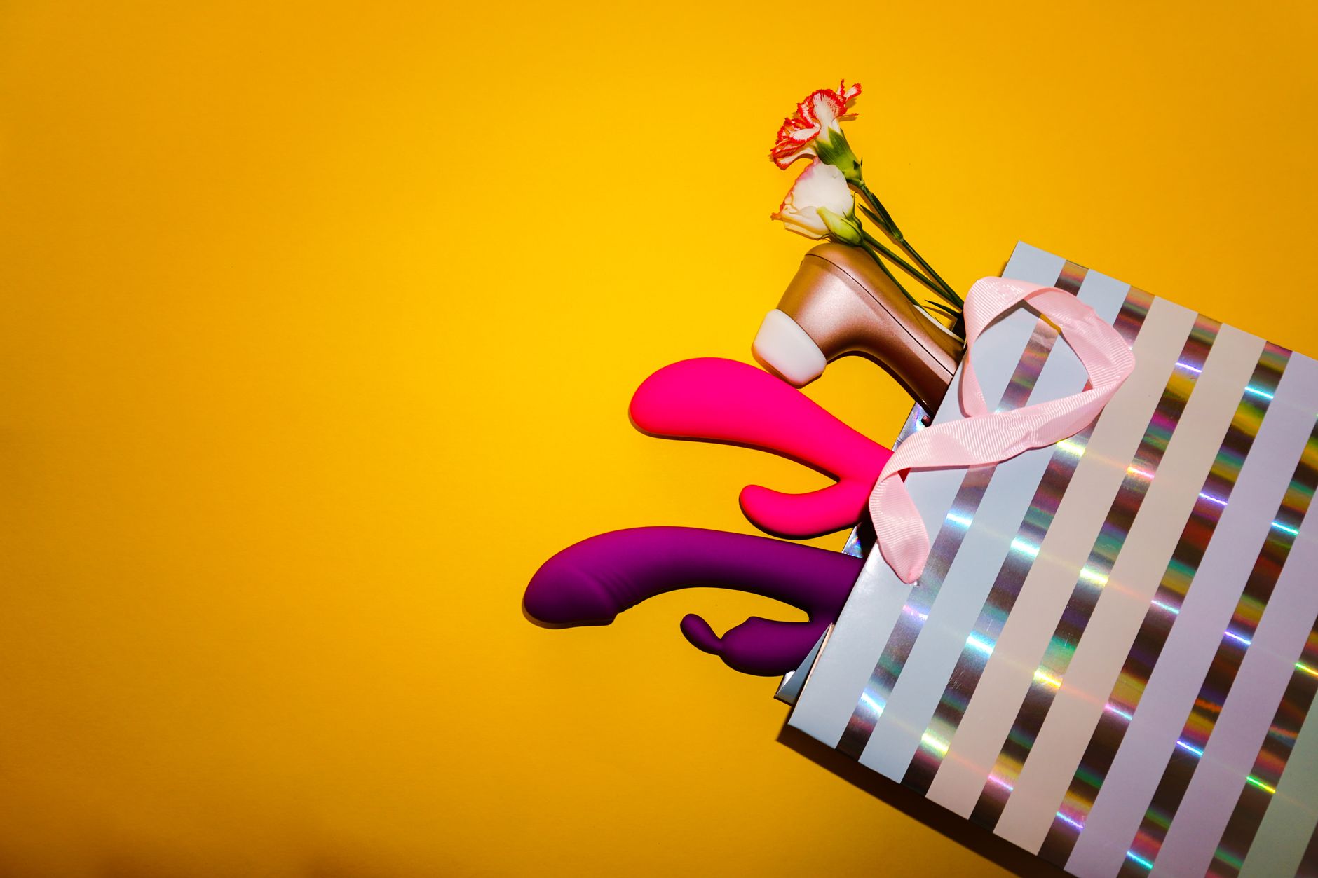 Vibromasseurs colorés avec fleurs dans un sac-cadeau holographique sur un fond jaune vif